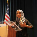 Omar SNUBS Senate — Why Risk Another Brawl? Woman speaking at podium with American flag background.