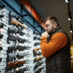 Standard Pistol Suddenly ILLEGAL—Virginia’s Stunning Move… Man contemplating firearms displayed on wall.