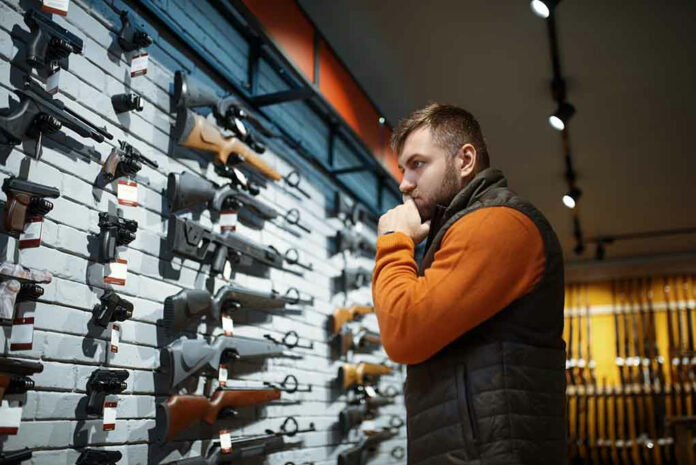 1686855364 Man contemplating firearms displayed on wall.