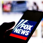 WILD ‘Mask’ Claims Shake Fox News Segment – VIDEO….. Silhouette hand holding phone displaying Fox News logo.