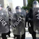 Police in riot gear holding shields in a formation.