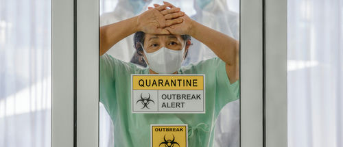 shutterstock_1648211998.jpg A healthcare worker in a green uniform and mask, looking distressed behind a glass door marked with quarantine signs