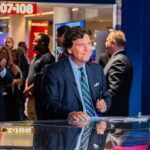 Tucker Carlson’s Holocaust Rhetoric SHOCKS America… News anchor smiling in a television studio with an audience in the background