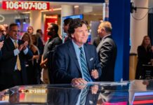 Tucker Carlson’s Holocaust Rhetoric SHOCKS America… News anchor smiling in a television studio with an audience in the background