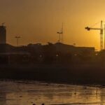 Air-Raid Chaos: Kuwait’s Terrifying Night City skyline with construction cranes at sunset by a reflective shoreline