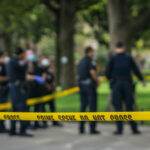 Carjacker Mows Down Ten — CHAOS! Police officers behind yellow crime scene tape in park.