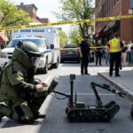 Explosive Find Triggers Mass Evacuations A bomb squad technician operating a robot in a street surrounded by police and emergency personnel