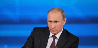 A serious-looking political figure seated at a press conference with a blue background