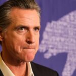 Federal Review Intensifies Pressure On Newsom A serious-looking political figure at a press event with a bear graphic in the background