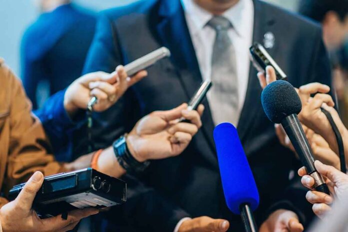 Journalists holding microphones interview a person in a suit.