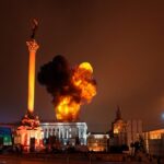 Large blaze in city square near tall monument at night.