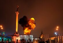 Large blaze in city square near tall monument at night.