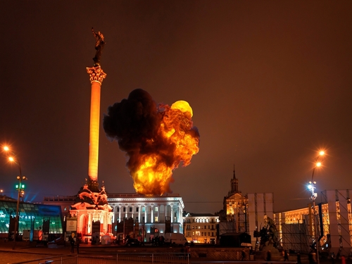 Large blaze in city square near tall monument at night.